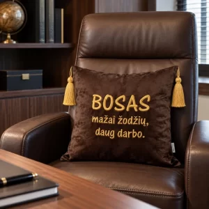 Tamsiai ruda dekoratyvinė pagalvėlė su aukso spalvos siuvinėtu užrašu „BOSAS – mažai žodžių, daug darbo“ ir kutais
