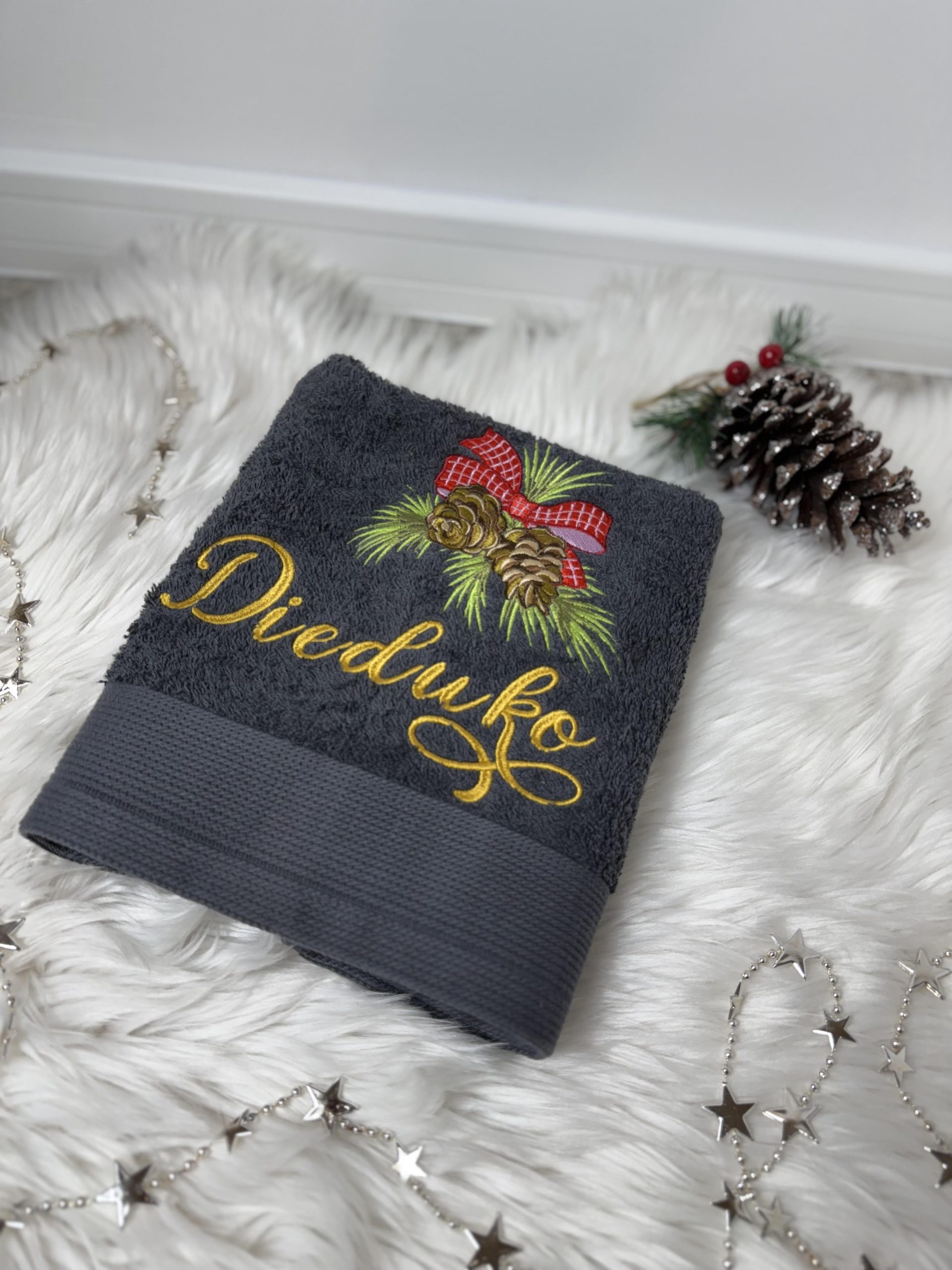 Pilkas siuvinėtas rankšluostis su užrašu „Dieduko“, puoštas eglės šakelėmis ir kankorėžiais.